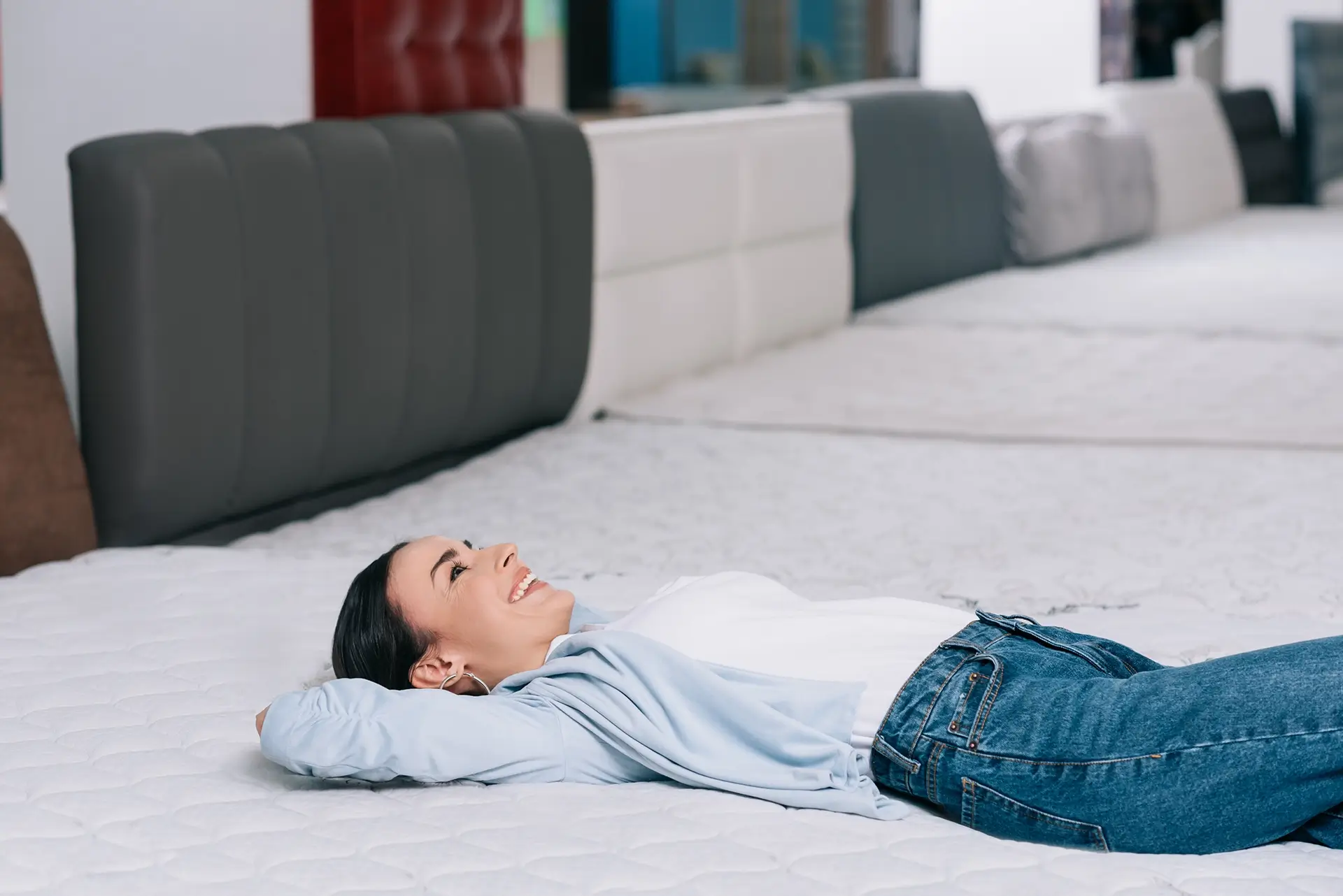 A woman in a pale blue overshirt smiles as she lies on a tester mattress in a store.