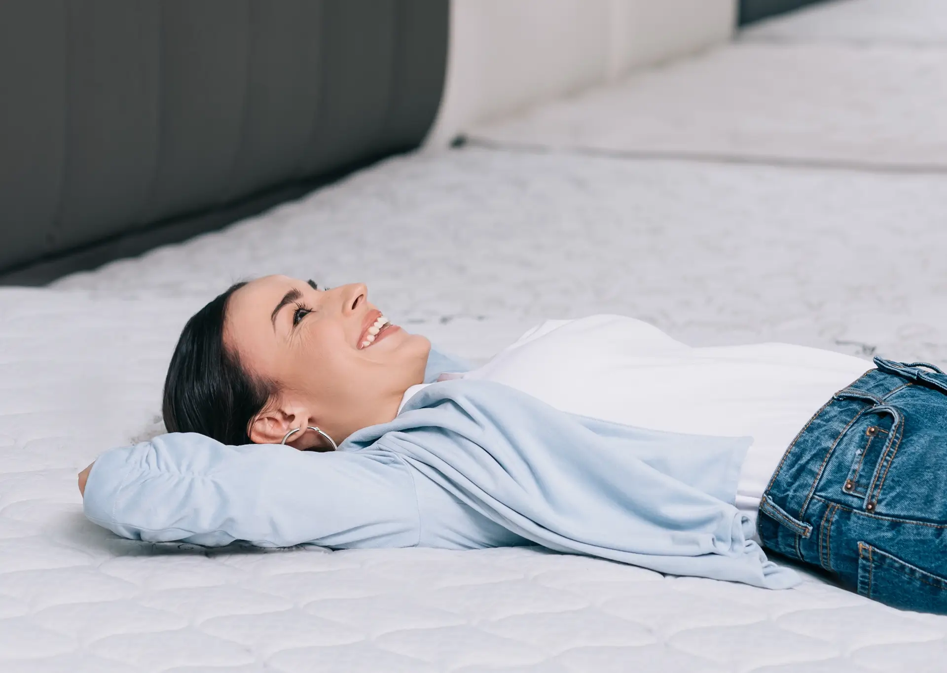 A woman in a pale blue overshirt smiles as she lies on a tester mattress in a store.