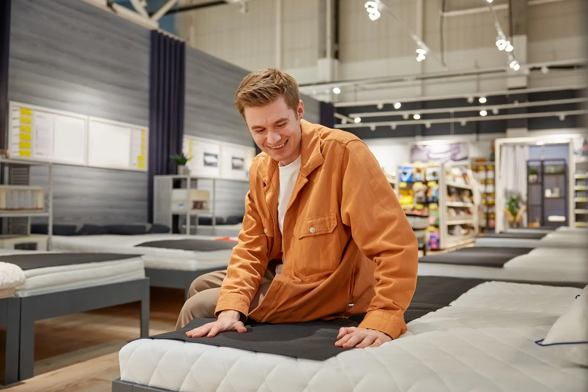 A young man in an orange overshirt sits on a tester bed at a store, feeling the mattress.
