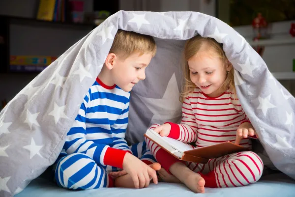 Two young siblings wearing pyjamas reading a book together with bedding over their heads.